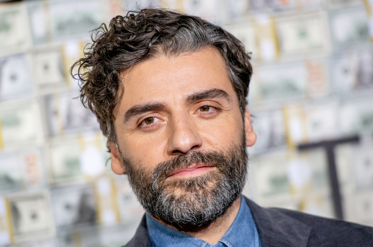 Oscar Isaac is wearing a suit and posing in front of a backdrop