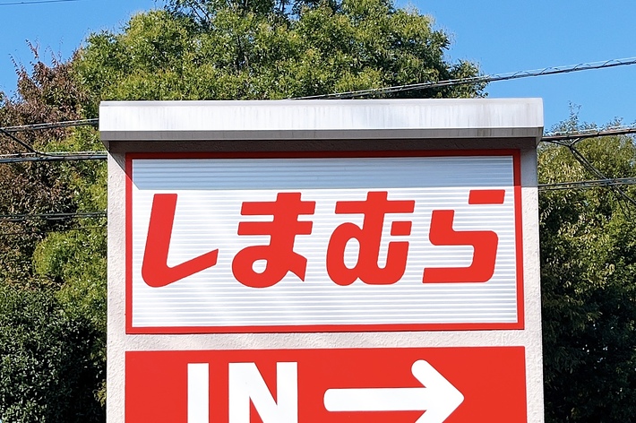 Large sign with Japanese text “しまむら” and an English “IN” with an arrow pointing right, indicating an entrance. Trees and a street in the background