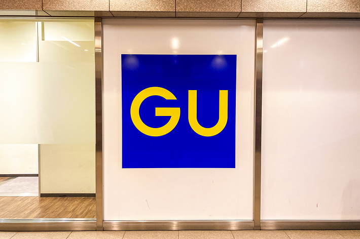 A store sign featuring the letters &ldquo;GU&rdquo; in yellow on a blue background. The sign is mounted on a white wall with a glass section to the left