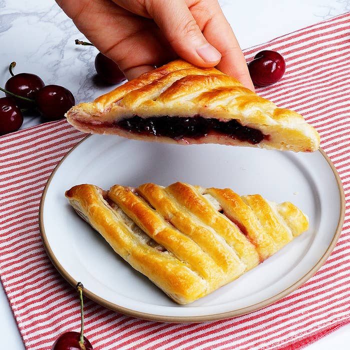 チェリーパイの一片を手で持ち上げている。お皿には他のチェリーパイの一片も載っている。背景にさくらんぼが見える。