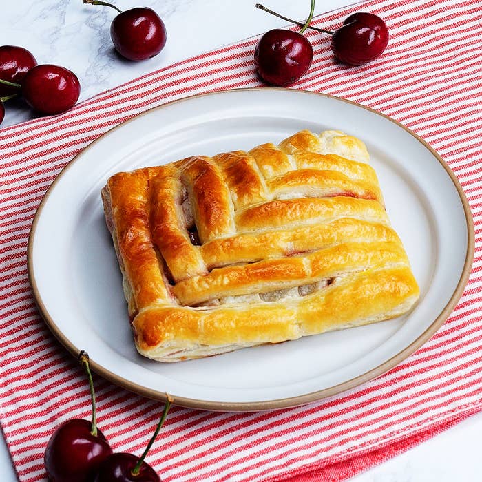 チェリーパイのデニッシュが白い皿に載せられ、赤のストライプのテーブルクロスと数個のチェリーが周りに配置されています。