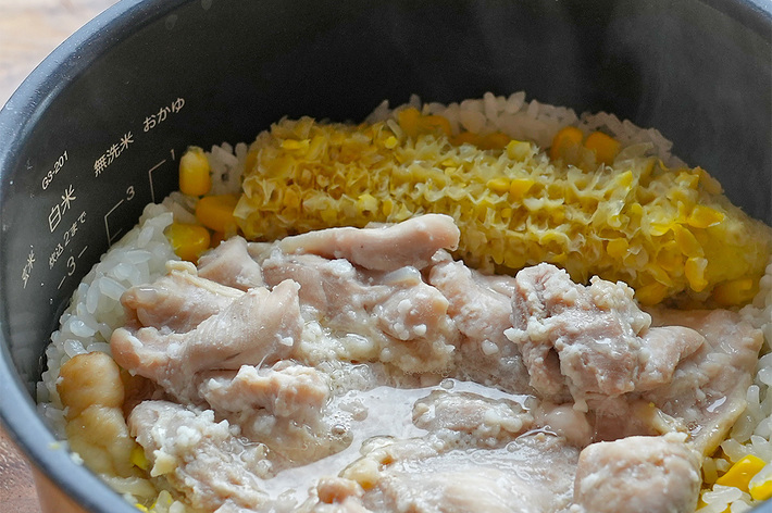 炊飯器の中で調理中のご飯、トウモロコシ、お肉の料理。