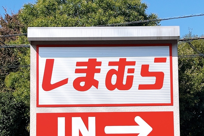 Sign at roadside entrance reads &ldquo;しまむら&rdquo; in Japanese, with &ldquo;IN →&rdquo; in English, directing vehicles to the right. Trees and road in the background