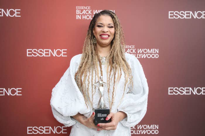Nkechi Okoro Carroll smiles while holding a trophy at the Essence Black Women in Hollywood event. She is wearing a white, elegant dress with puffed sleeves