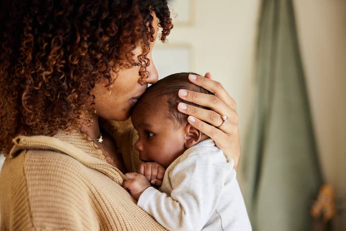 A person gently kisses the forehead of a baby they are holding close to their chest. The person wears a soft-texture sweater and a ring on their finger