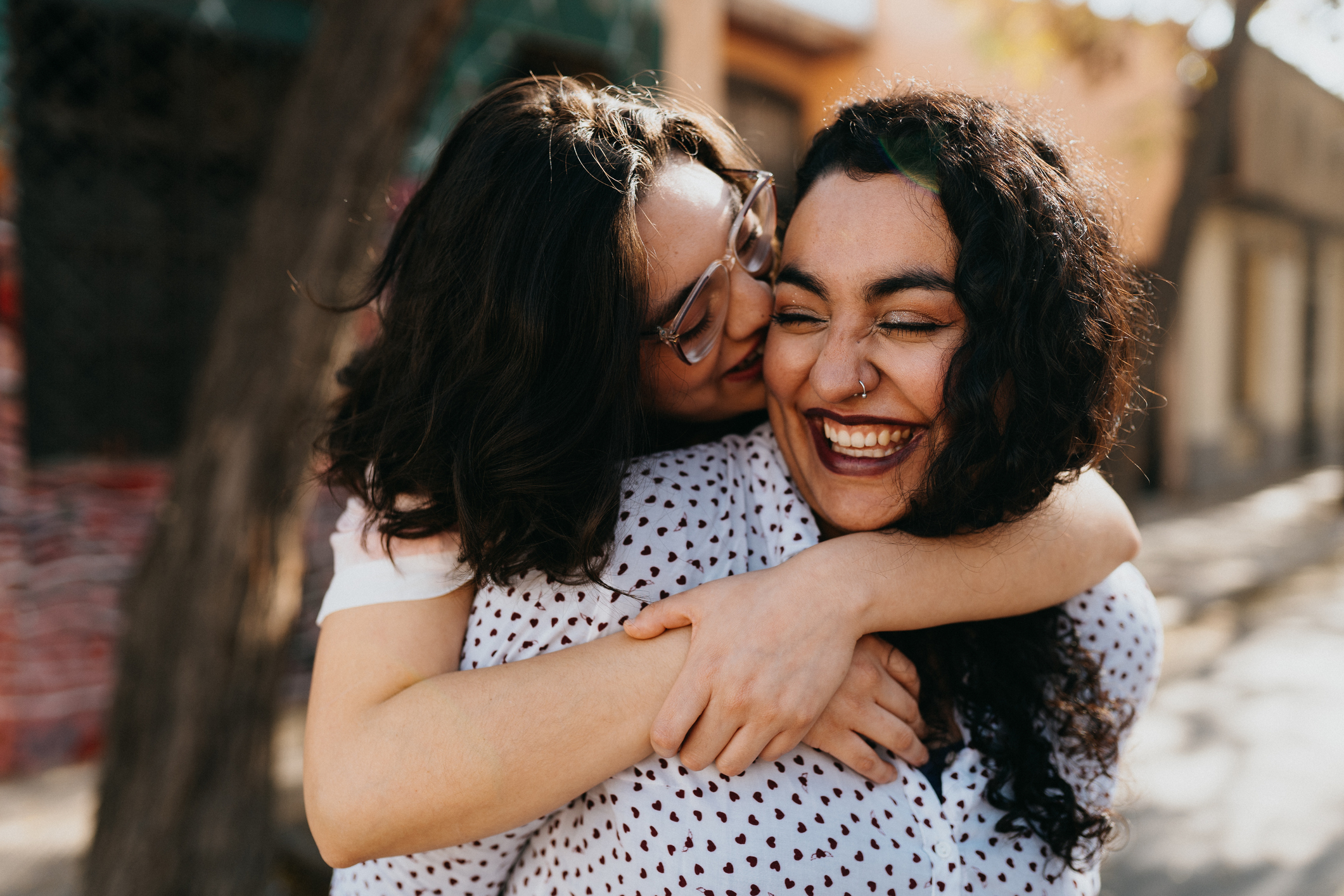 A person with curly hair smiles wide as they receive a warm hug from another person with glasses, both appear happy in an outdoor setting