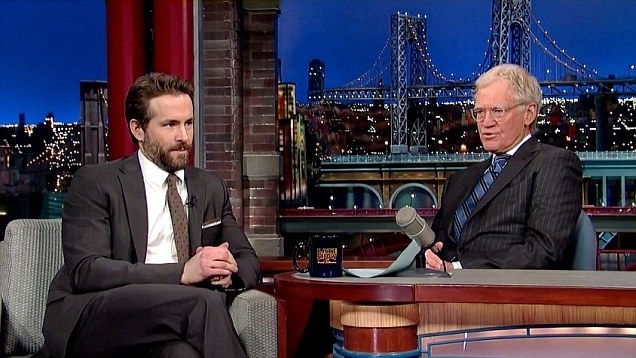 Ryan Reynolds, in a suit and tie, is being interviewed by David Letterman on his talk show set, with cityscape and bridge in the background
