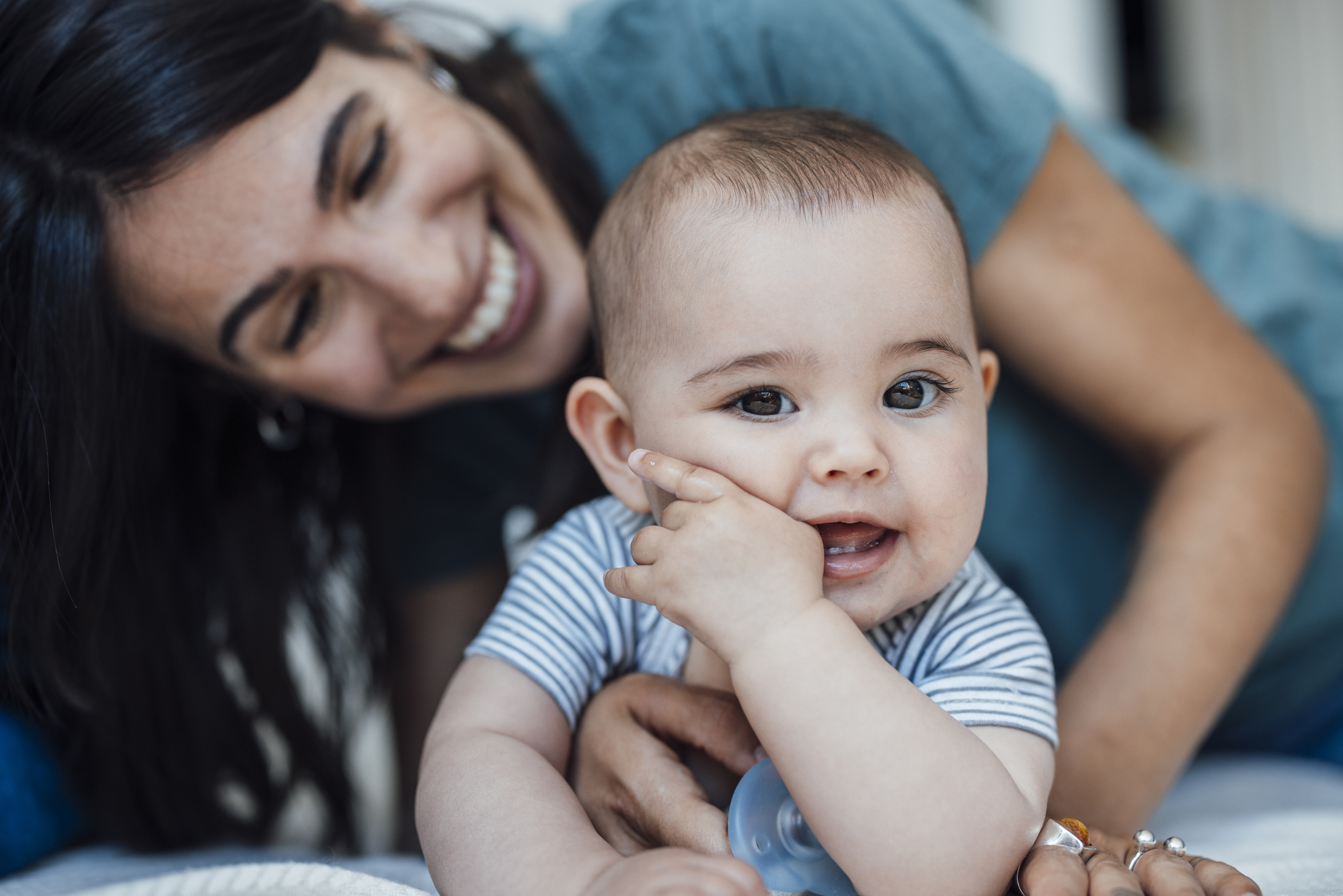 A woman smiles while lying down with an adorable baby, who is touching their face and looking at the camera