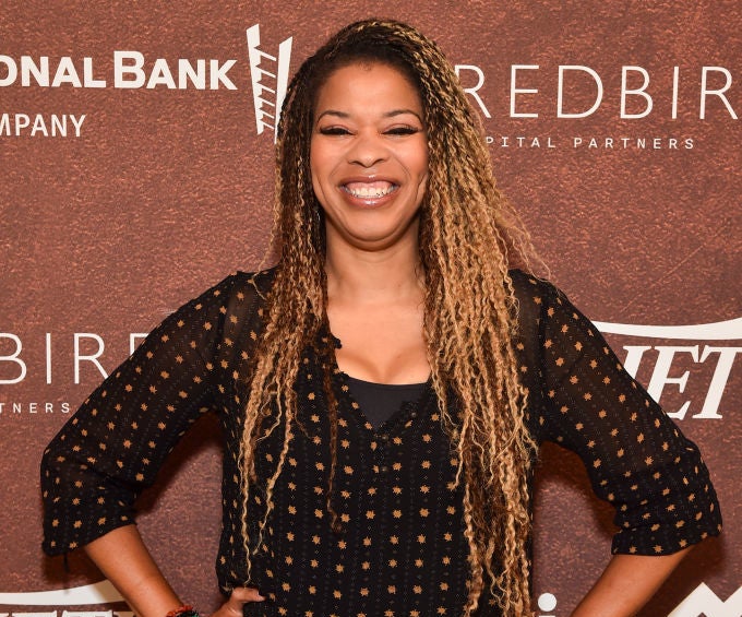 Nkechi smiles broadly, standing with hands on hips, wearing a black blouse and dark jeans, at a Variety-sponsored event