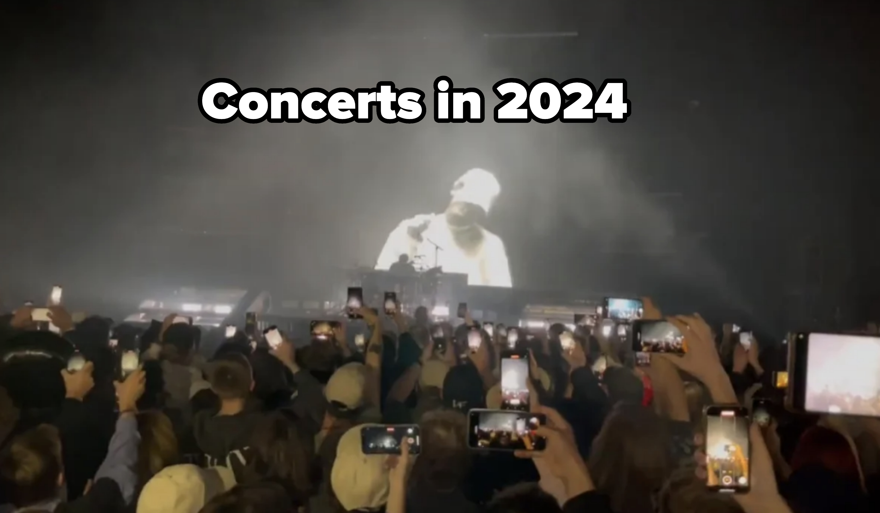 A large audience holds up phones to capture rapper Ye performing on stage in a dimly lit, smoky atmosphere