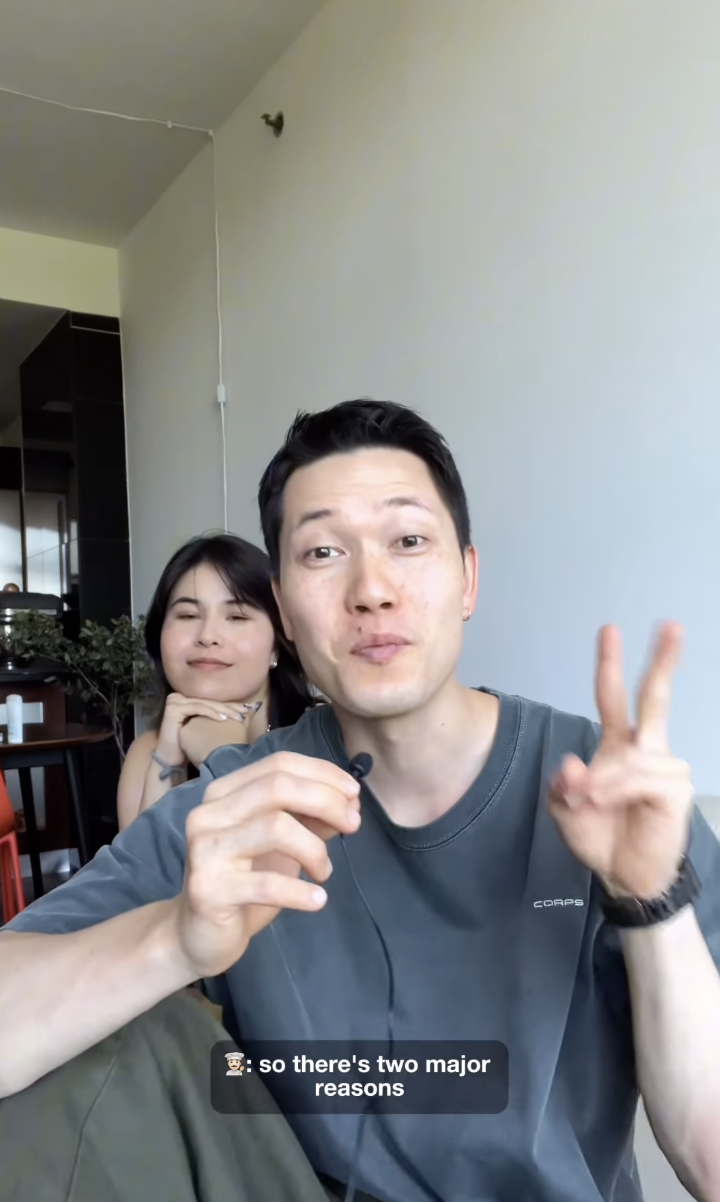 A man and woman sit casually indoors. The man speaks into a microphone and gestures with two fingers. Subtitles say: "so there's two major reasons"