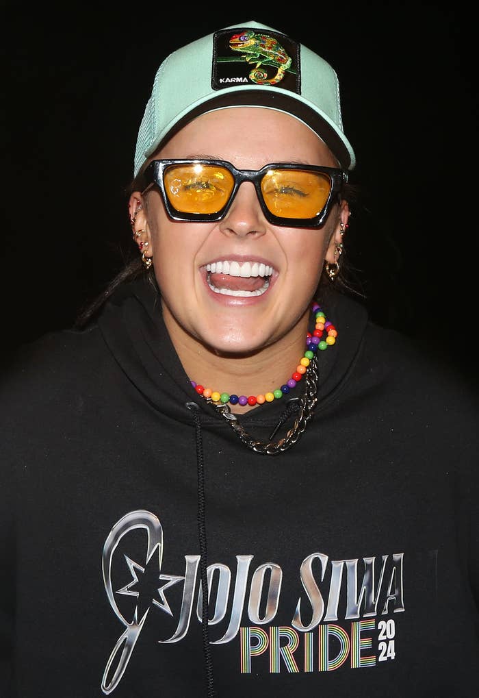 JoJo Siwa wears a cap, orange-tinted glasses, a colorful bead necklace, and a hoodie with &quot;JoJo Siwa Pride 2024&quot; printed on it. She is smiling widely