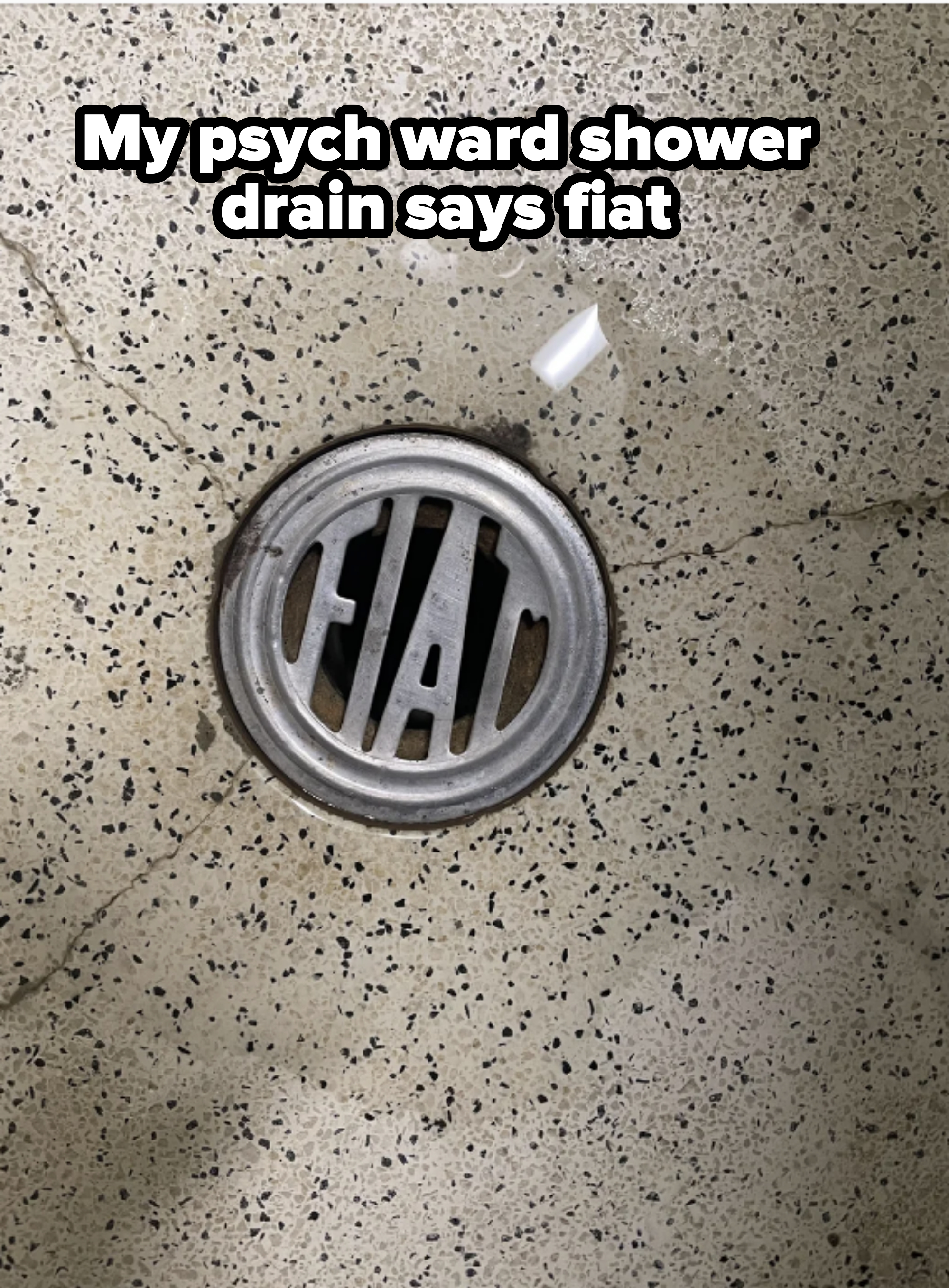 Metal floor drain on a speckled surface, with a small piece of trash nearby