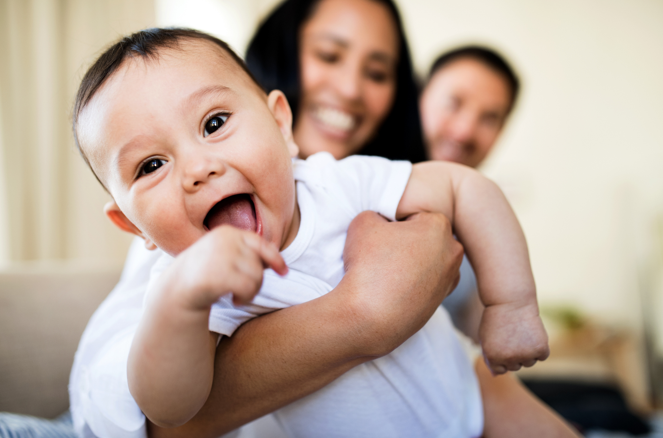 Smiling baby in a white onesie is held by an adult. Two adults are smiling in the background, slightly out of focus. Names are not known
