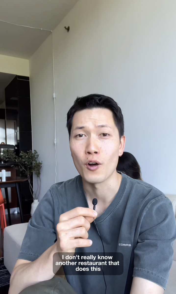 Man holding a microphone, speaking. Caption reads: "I don't really know another restaurant that does this." A plant is seen in the background