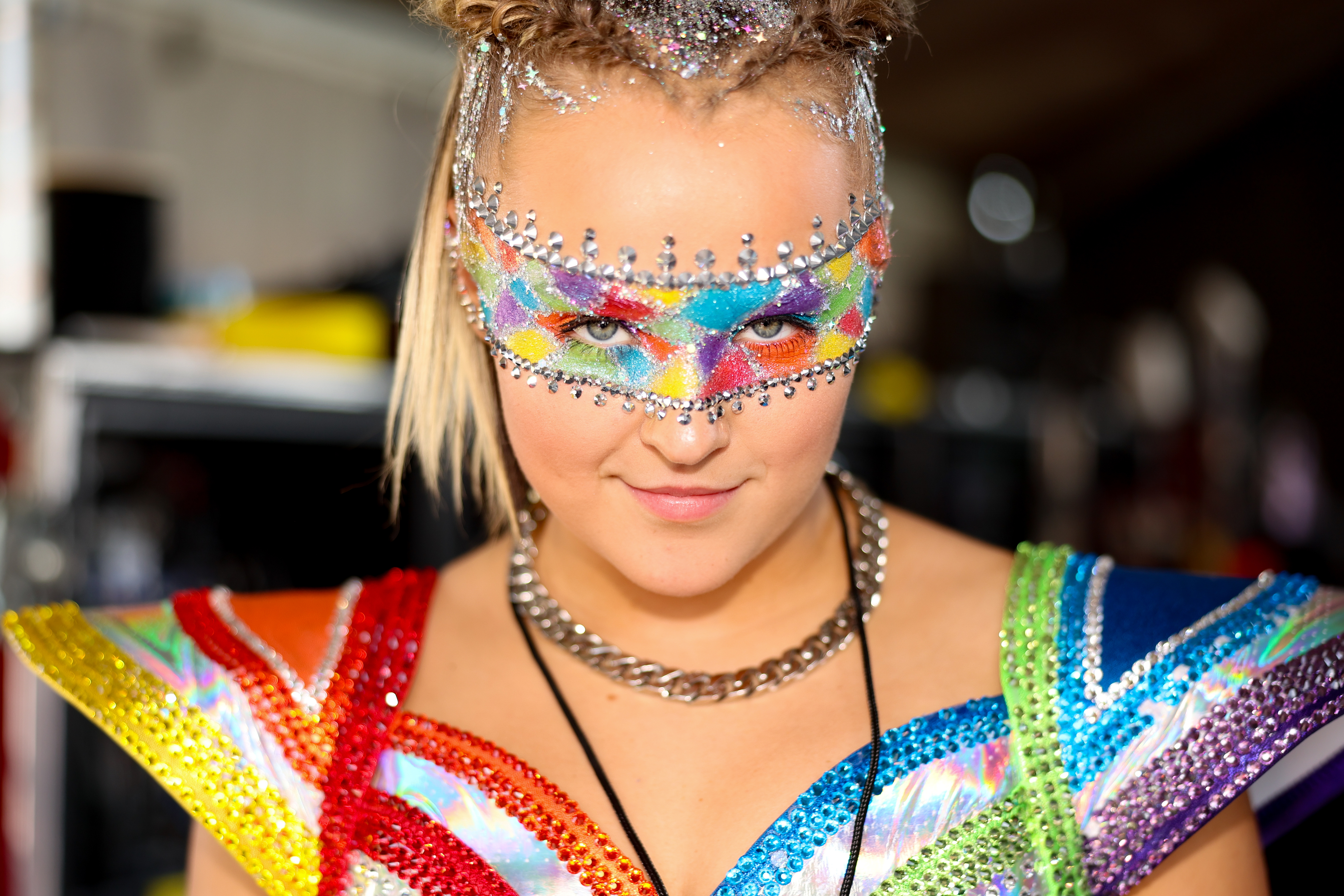 Jojo Siwa in a vibrant, bejeweled outfit with a colorful, glittering face mask and braided hair, posing for the camera
