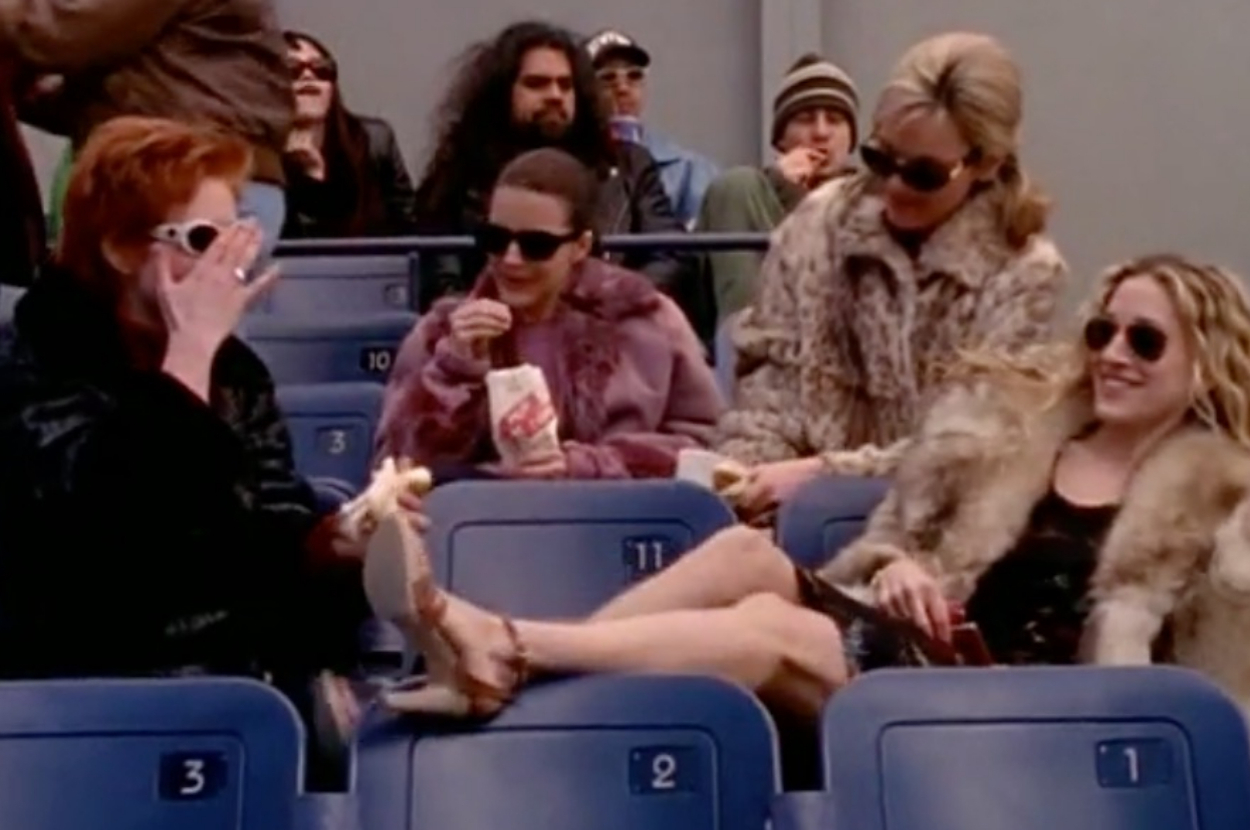 Kim Cattrall, Kristin Davis, Sarah Jessica Parker, and Cynthia Nixon sitting in stadium seats, dressed in stylish casual outfits and sunglasses, sharing a light moment