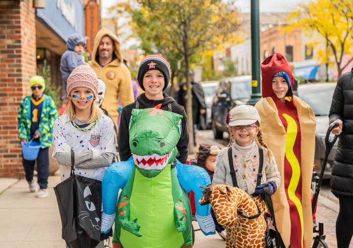 Kids in costume, including a dinosaur, hot dog, animal onesies, and casual sporty outfits, walking on a sidewalk during a Halloween event