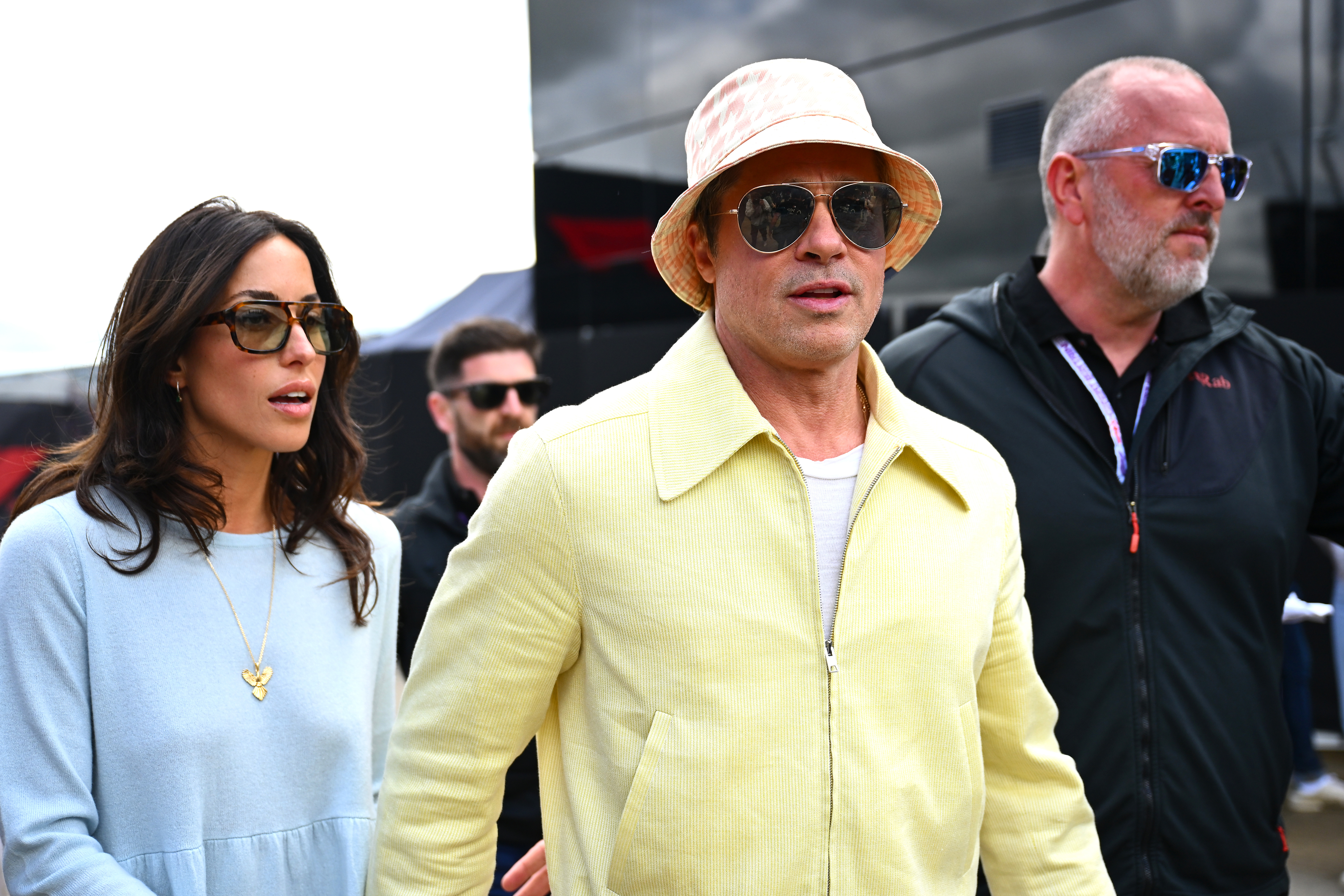 Brad Pitt wearing a jacket and a bucket hat walks beside Ines de Ramon, dressed in a light outfit, both accompanied by a bodyguard outdoors