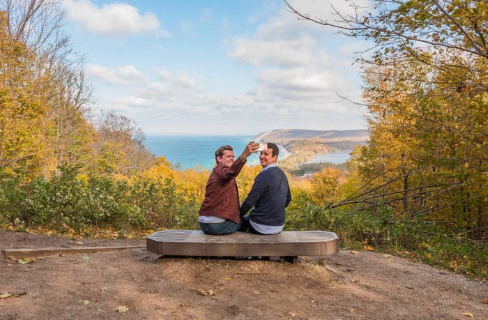 Two people sit on a bench overlooking a scenic view of mountains, forests, and the ocean. One person is showing something to the other