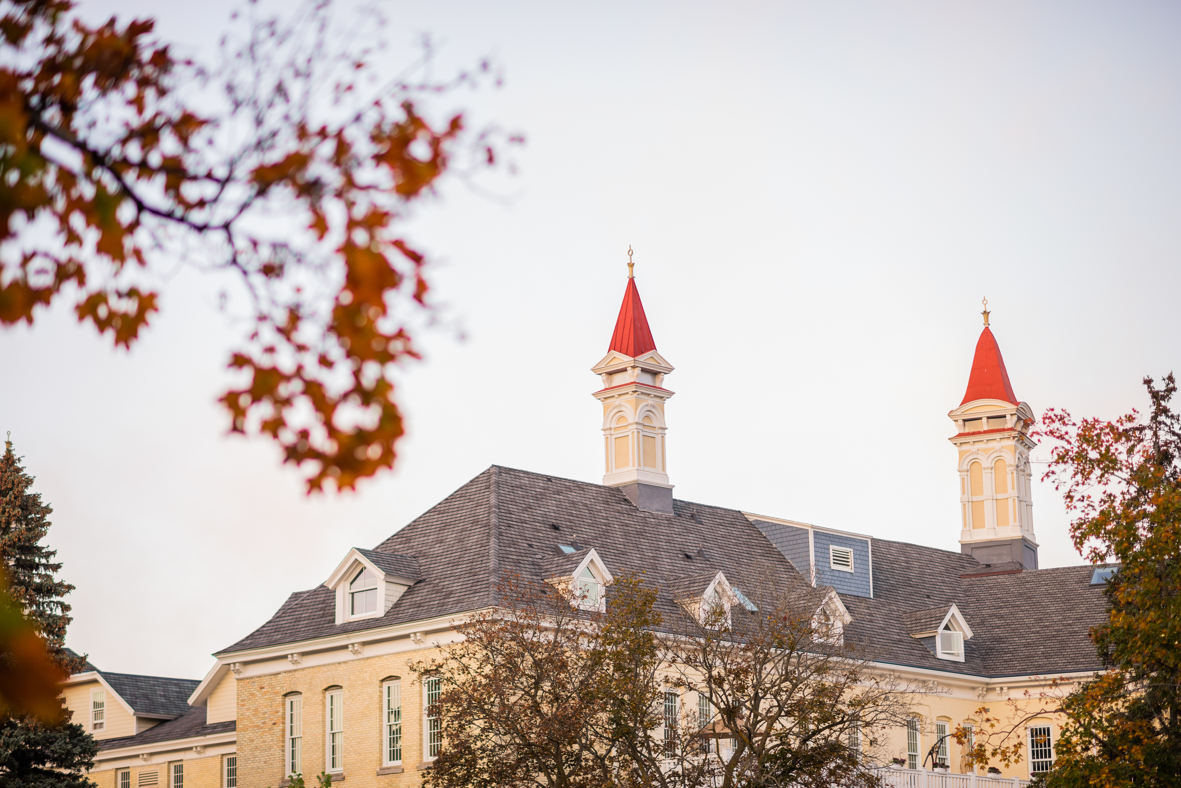 Exterior view of a large, historic building with two red-topped towers, surrounded by autumn trees