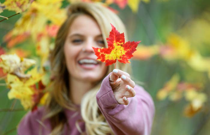 A smiling woman in a sweater holds a maple leaf close to the camera, with autumn foliage in the background