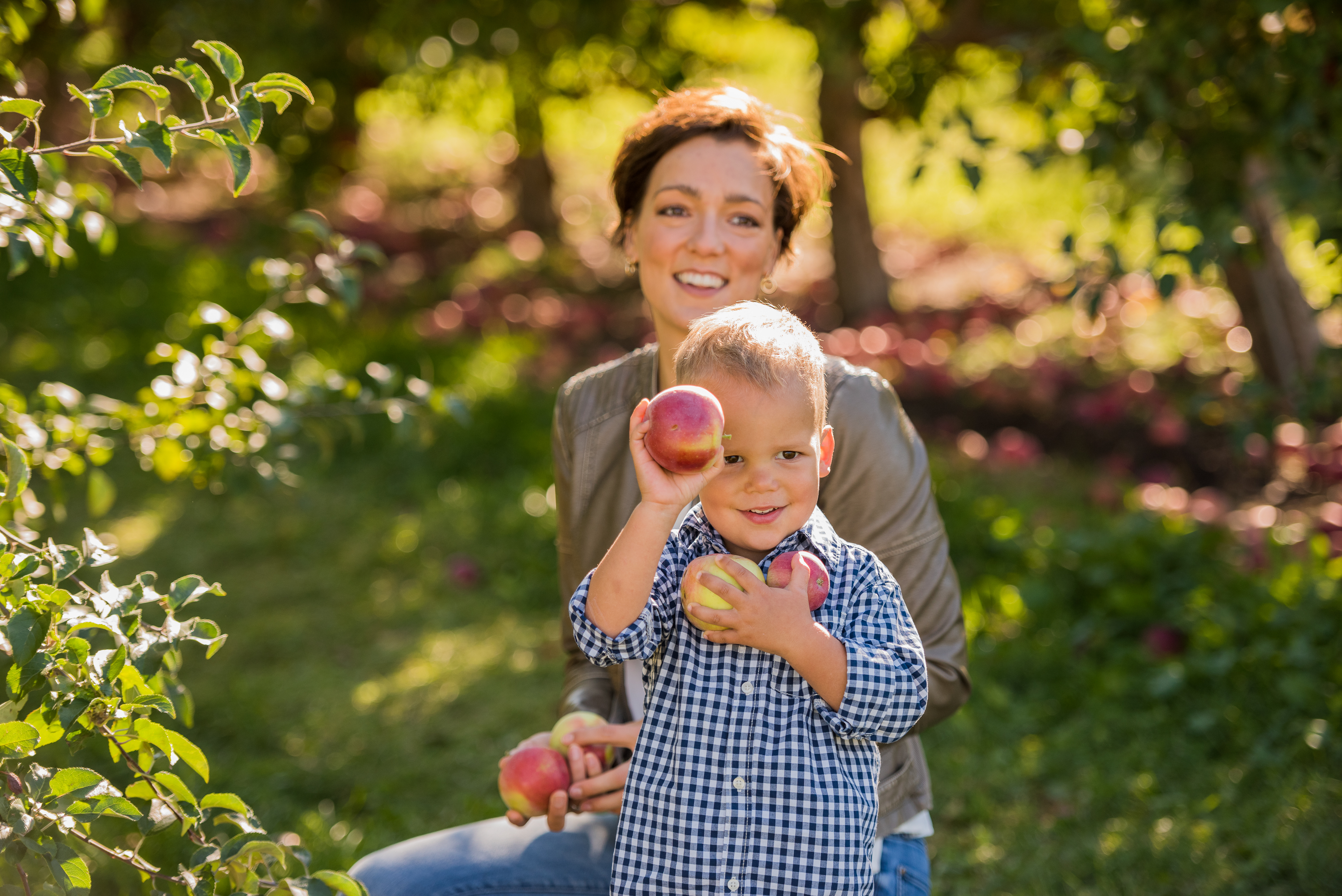 A woman smiles while sitting outdoors, holding peaches with a young boy who playfully holds a peach to his eye. Both appear to enjoy a sunny day in an orchard