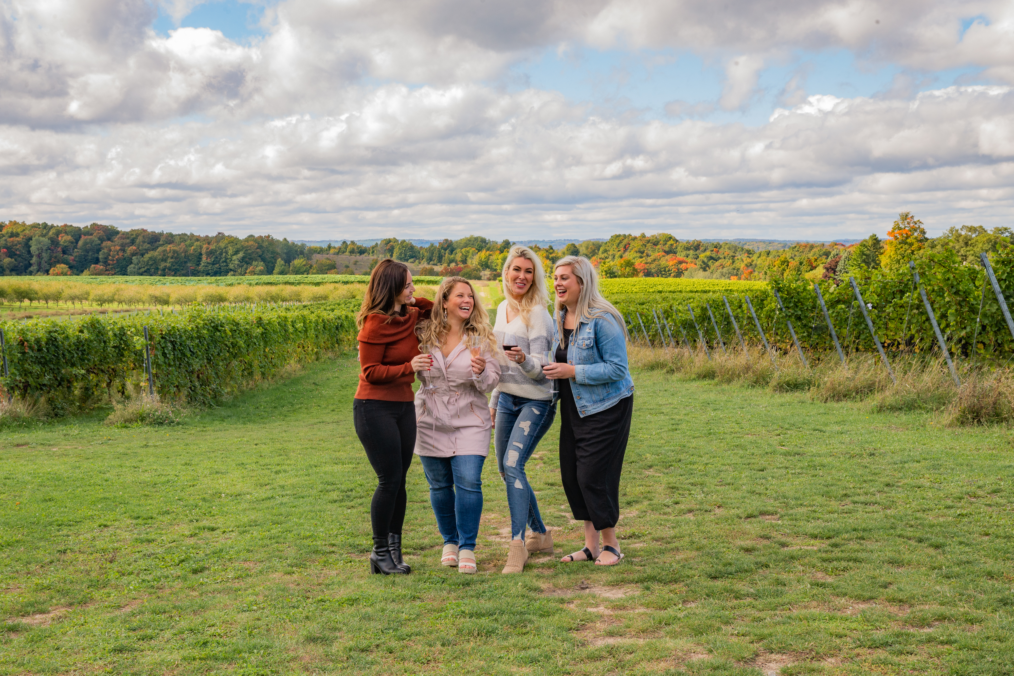Four women, standing in a vineyard on a cloudy day, share a joyful moment