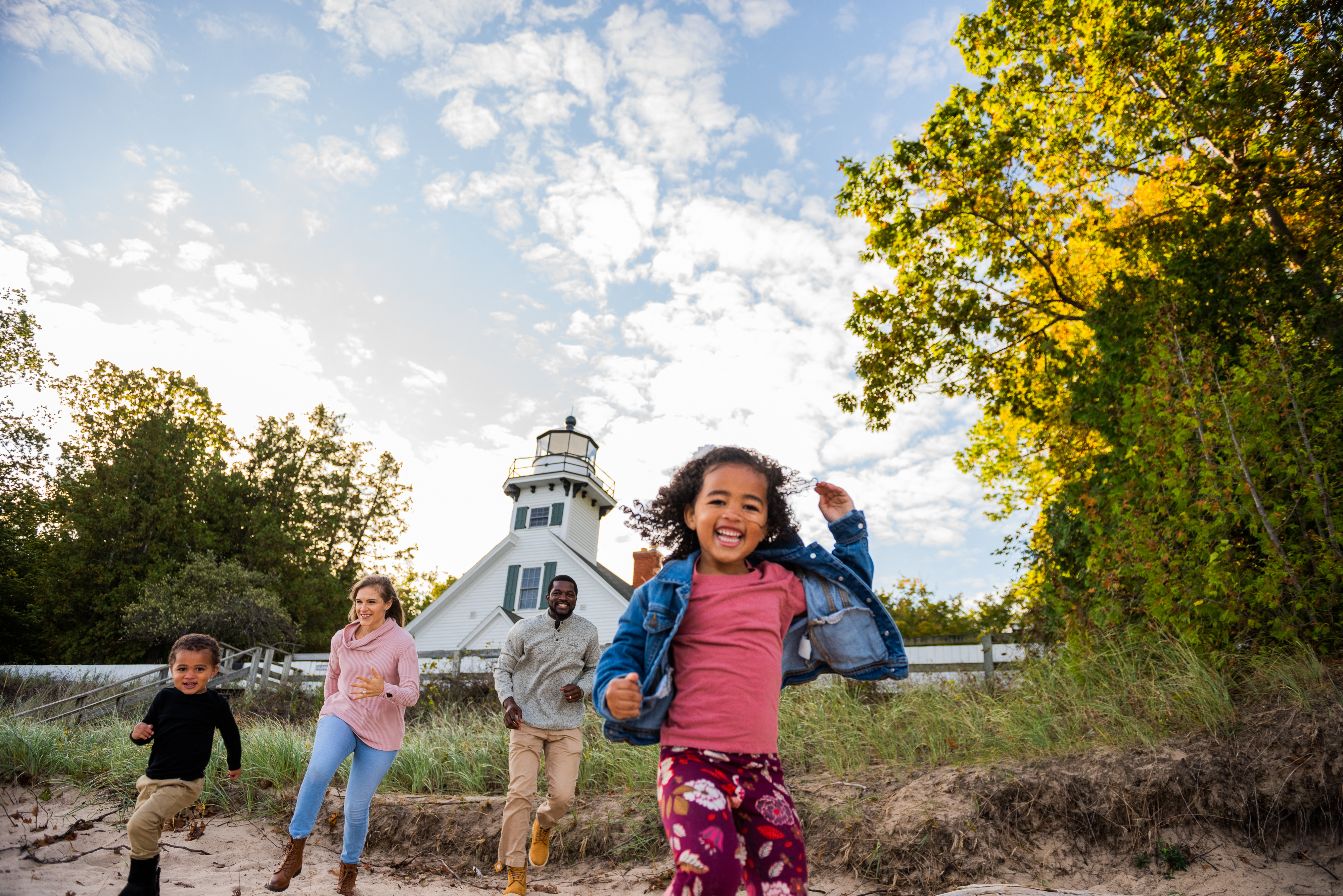 A family of four, two adults and two children, happily running on a beach near a lighthouse surrounded by trees