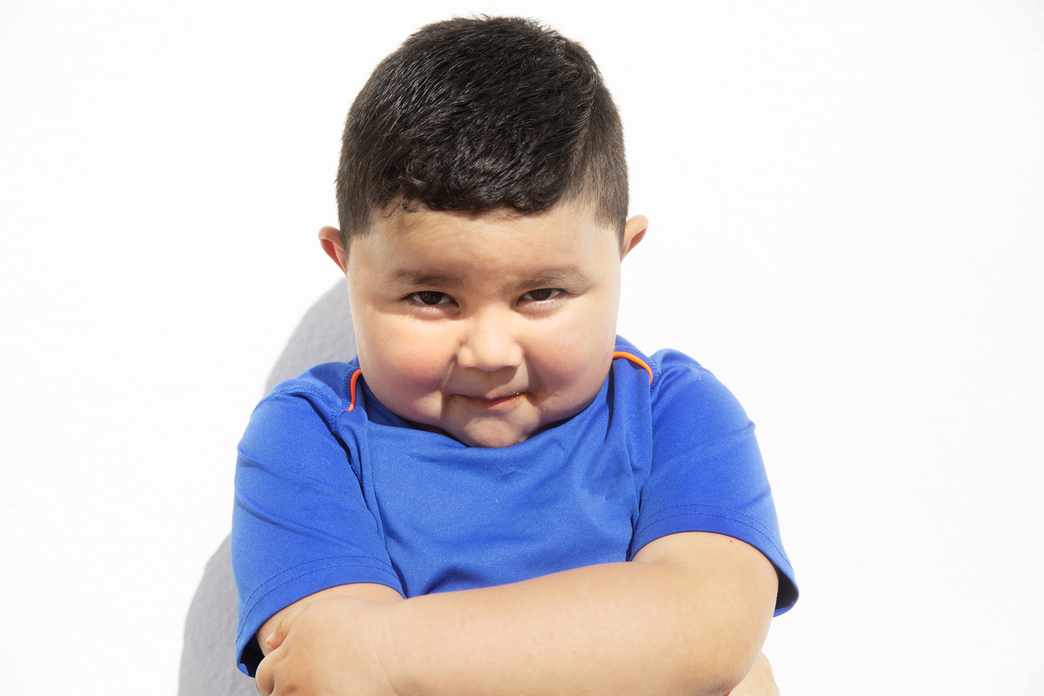 Young child with a mischievous expression, standing against a plain background, arms crossed wearing a short-sleeved shirt
