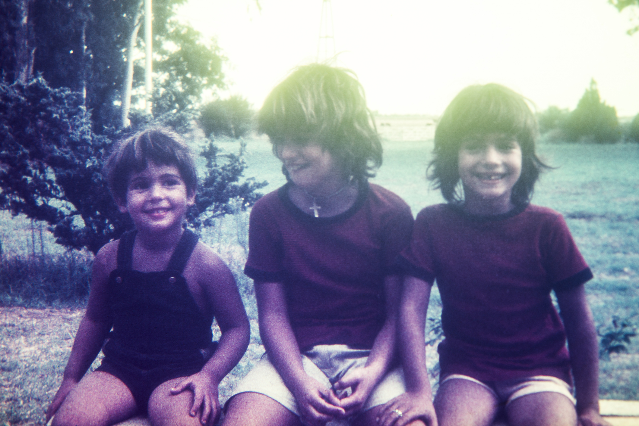 Three children sit outdoors, smiling at the camera. They appear to be enjoying a candid moment. The child on the left wears overalls, and the other two wear T-shirts