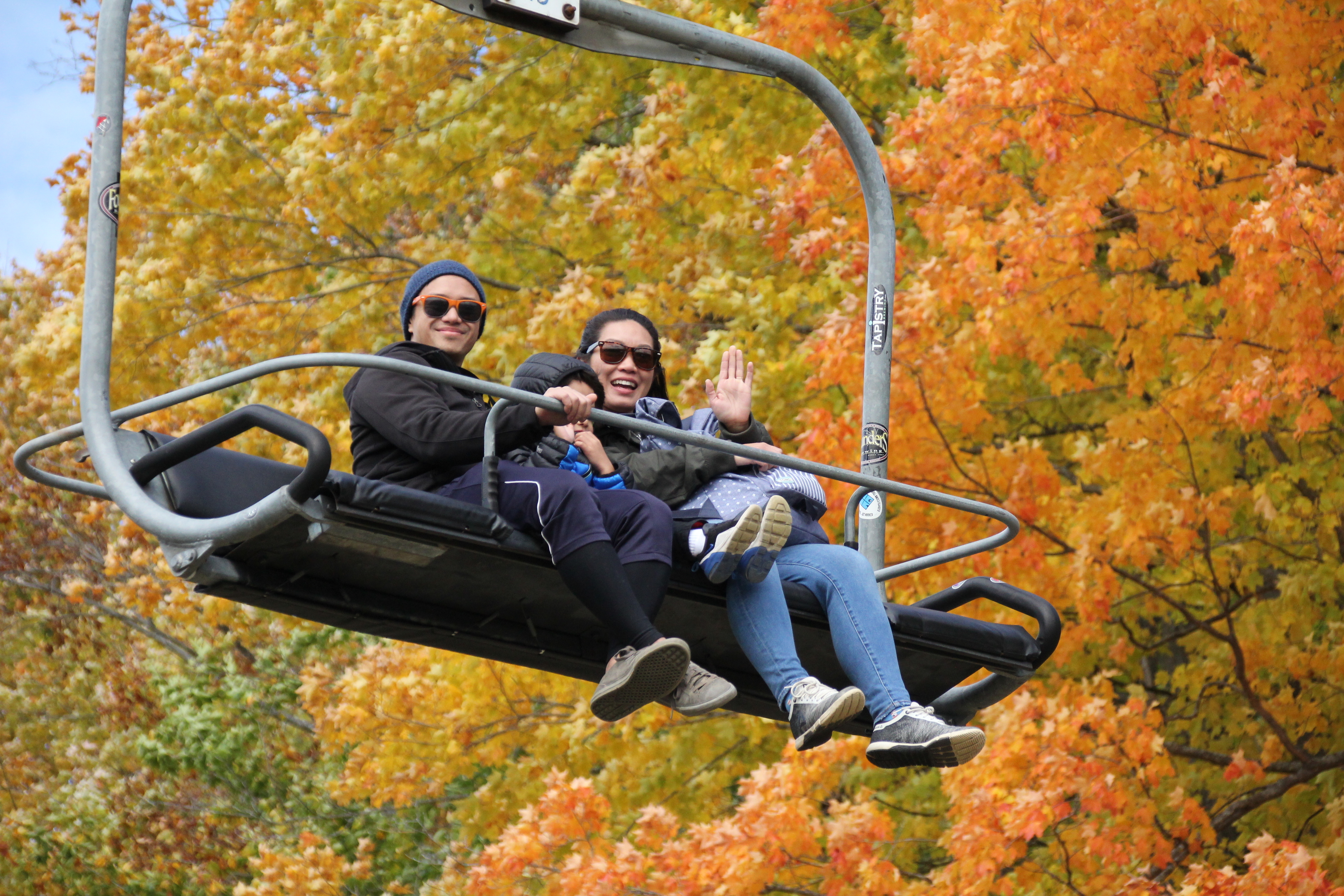 A man, woman, and children riding a chairlift in a fall setting with colorful autumn trees in the background; the smiling woman waves at the camera