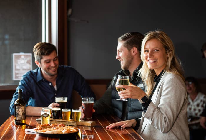 Three people enjoying drinks and pizza at a bar. The woman in the foreground wearing a blazer smiles at the camera while holding a glass of white wine