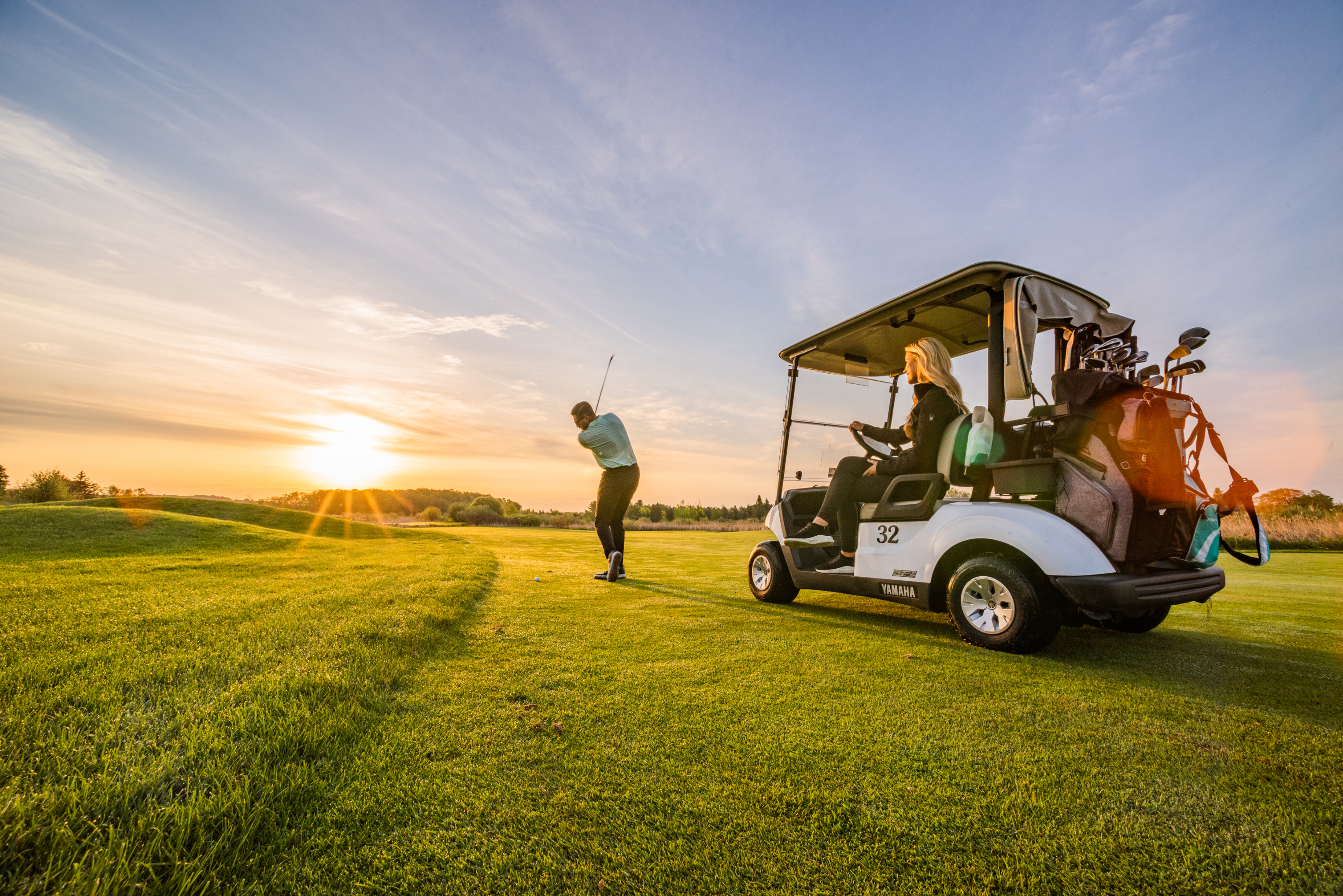 Two people golfing at sunset with one person swinging a golf club and another person seated in a golf cart numbered 32