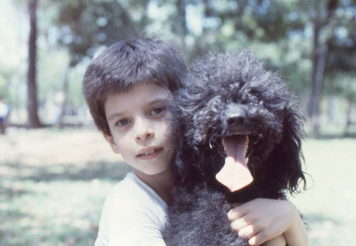 A young boy hugs a black poodle dog outdoors, both facing the camera. Trees and greenery are visible in the background