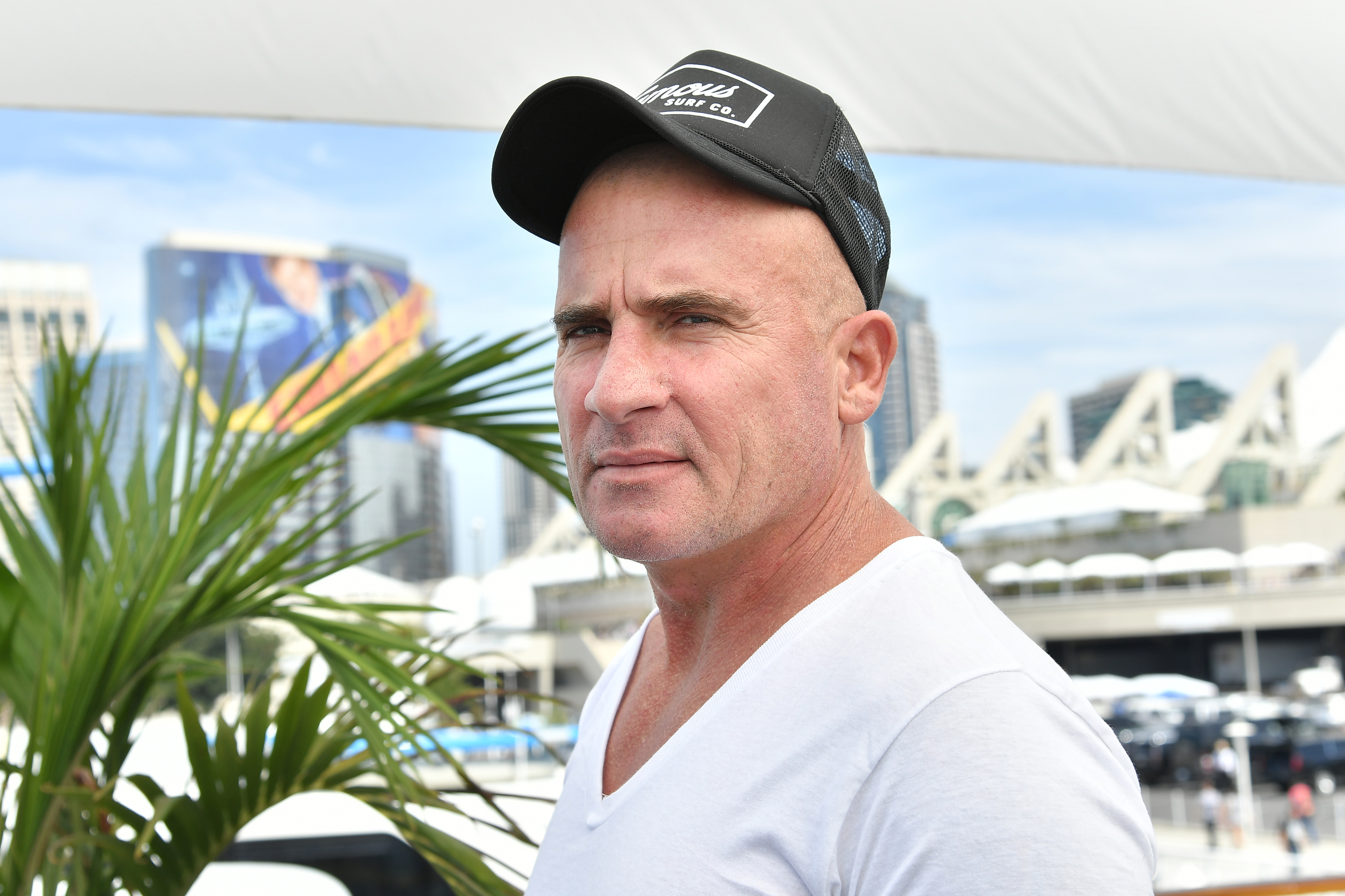 Dominic Purcell, in a casual outfit wearing a cap and a white shirt, stands outdoors with a cityscape in the background