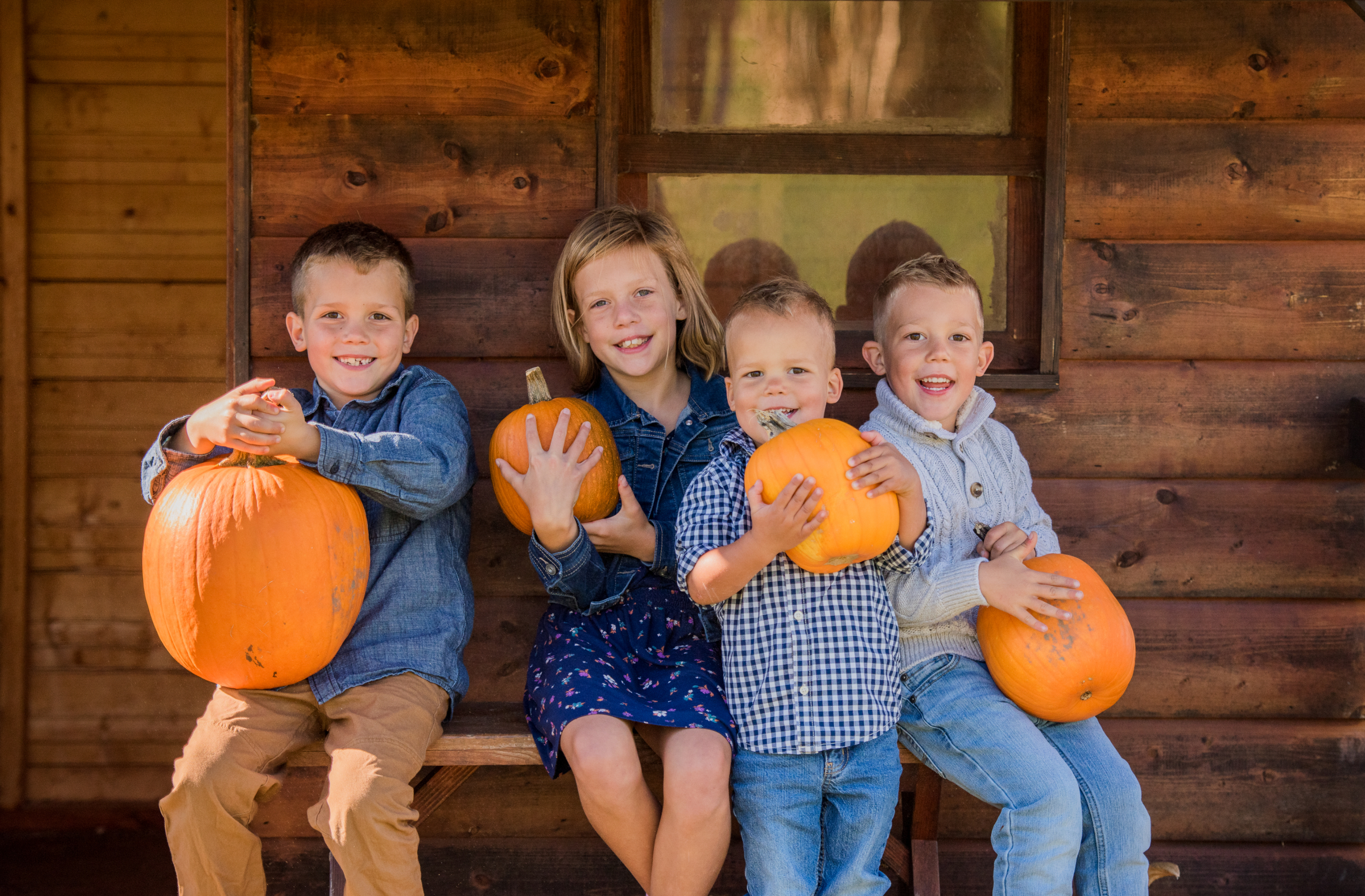 Four smiling children hold large pumpkins while sitting on a wooden bench in front of a rustic cabin