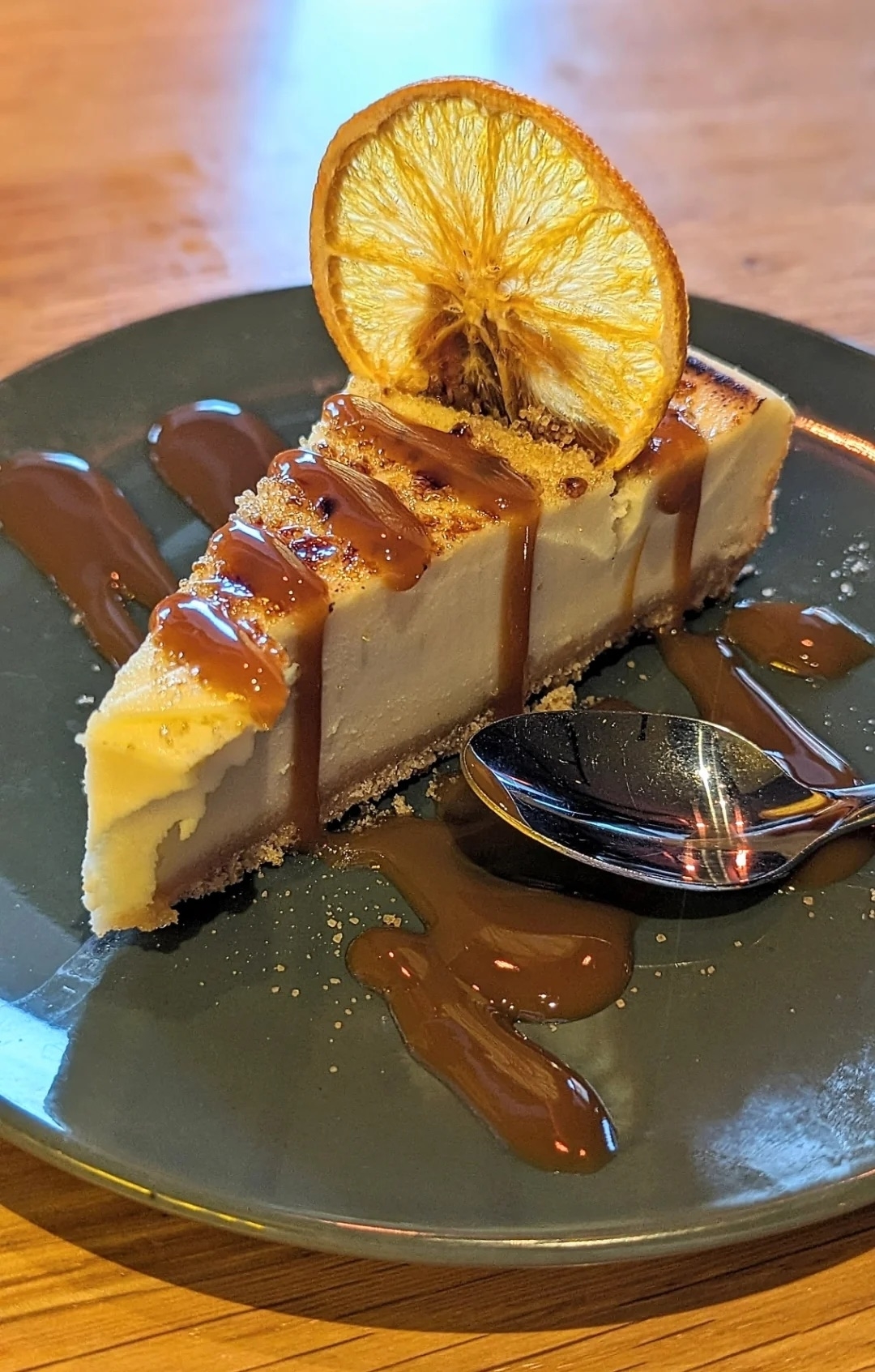 A slice of cheesecake garnished with a dried orange slice and caramel drizzle, served on a blue plate with a spoon