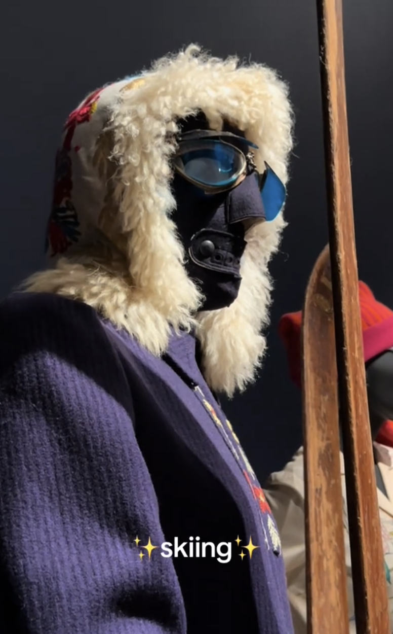 A person wearing a fur-lined hood, ski goggles, and holding vintage wooden skis, with the caption &quot;skiing&quot; at the bottom