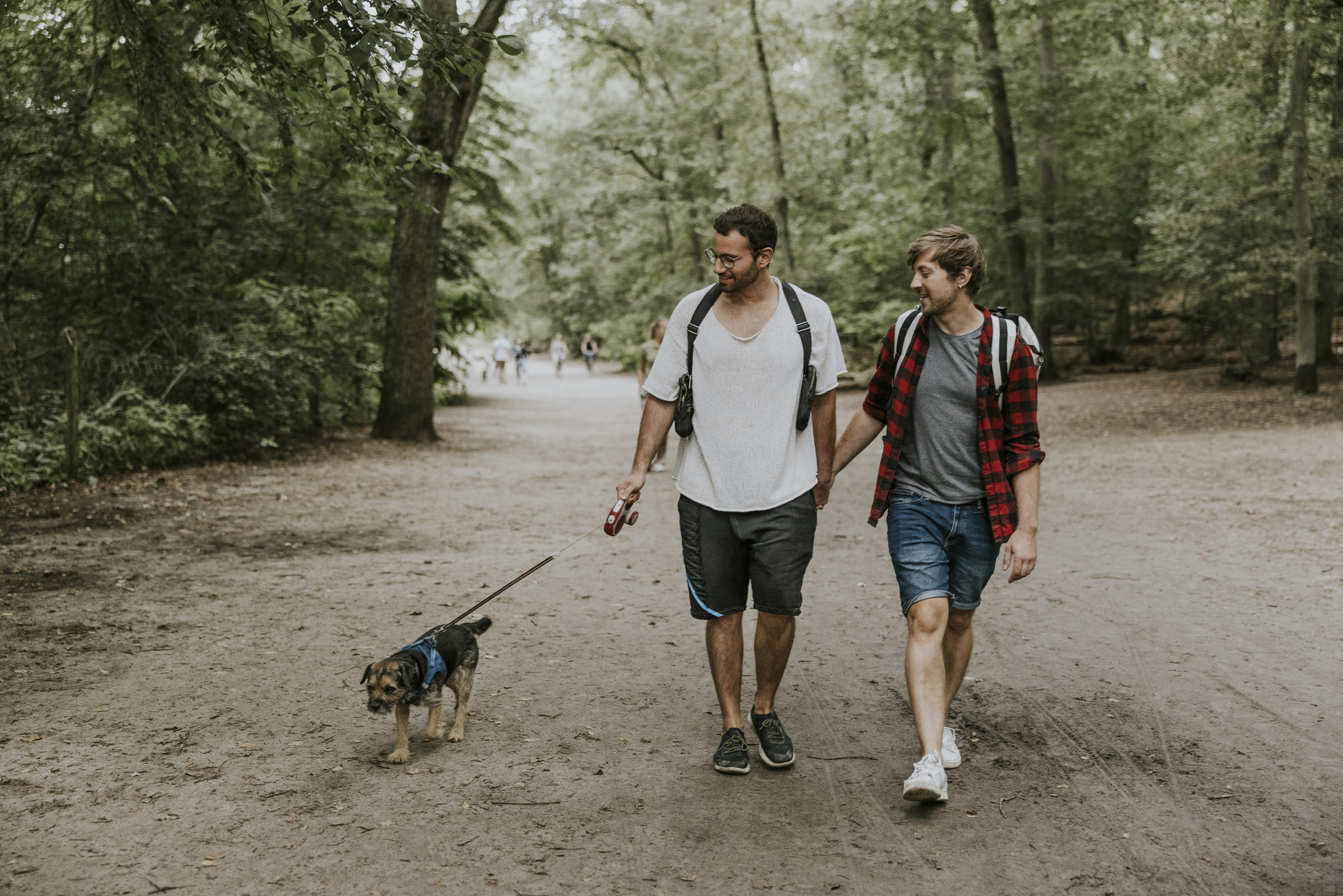 Two men, one in a white shirt and the other in a red checkered shirt, walk a small dog in a park. Both carry backpacks and appear to be enjoying the walk