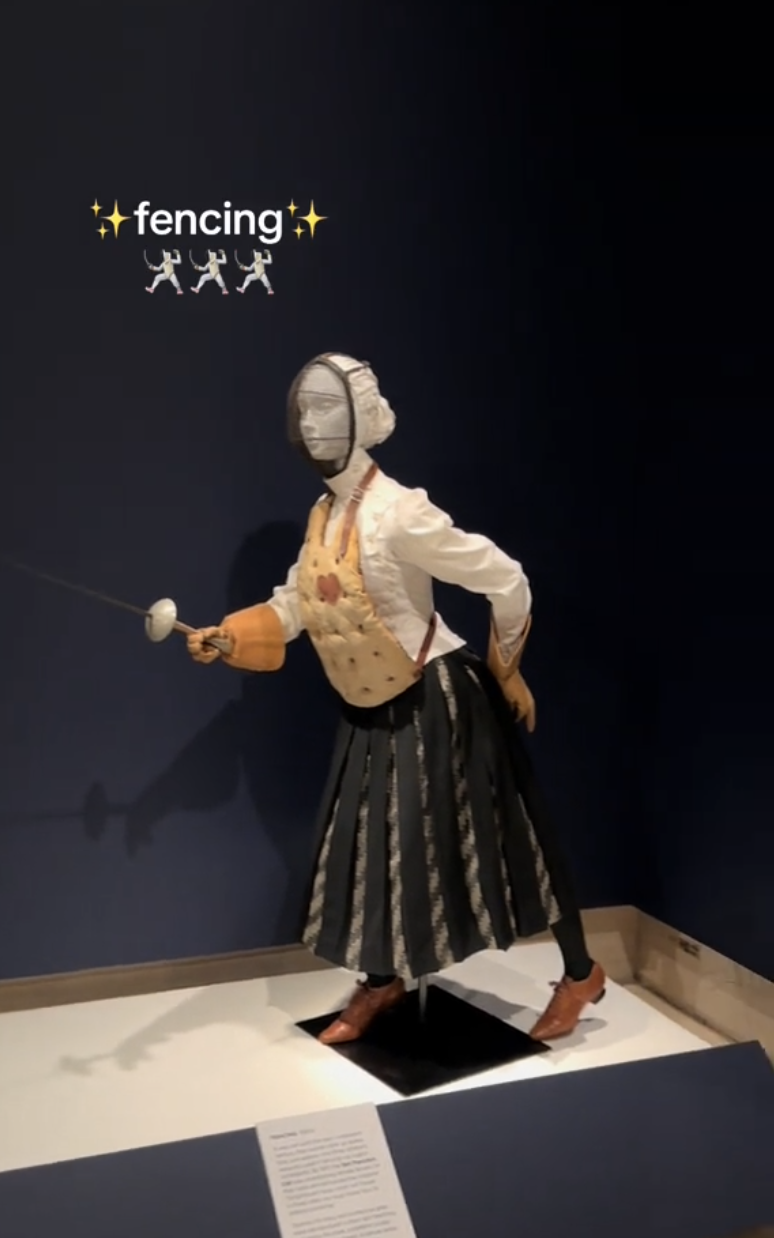 A museum display of a historical fencing outfit on a mannequin, complete with a mask and sword, under the caption &quot;fencing&quot; with fencing emojis