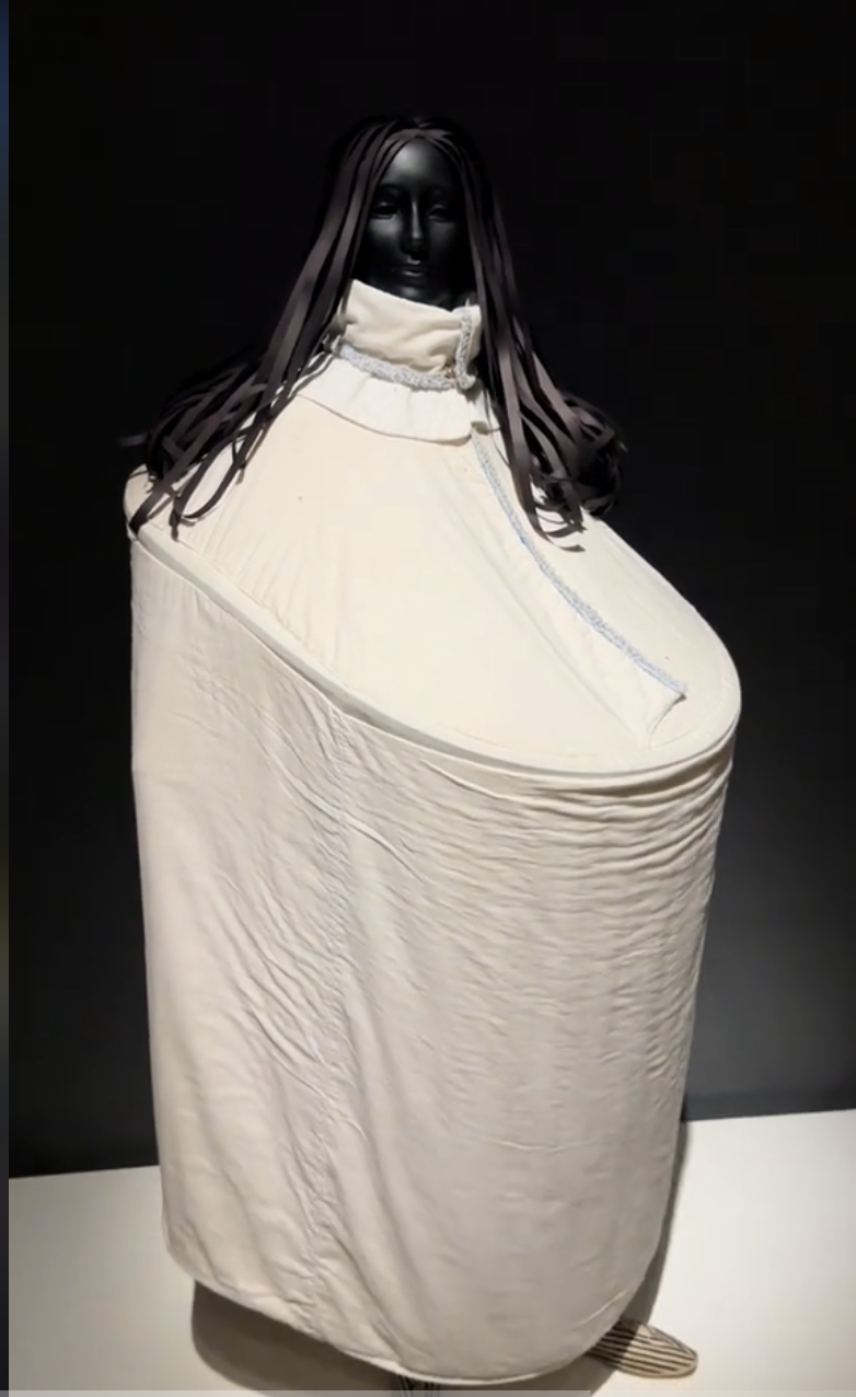 Person in an avant-garde, sculptural white outfit with a high collar and black face covering. The outfit resembles a large, padded cylindrical shape