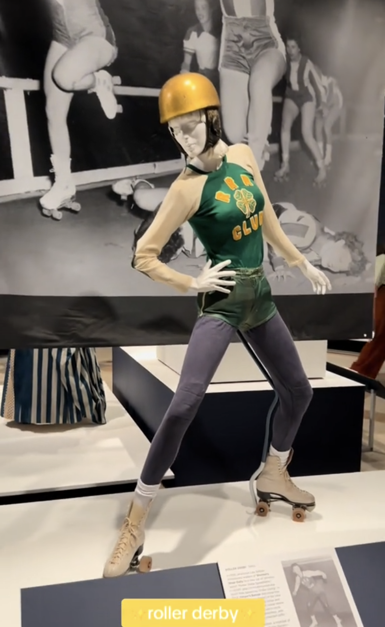 Mannequin wearing a Gold Club roller derby outfit, including a golden helmet and roller skates, with a historical black and white photograph in the background