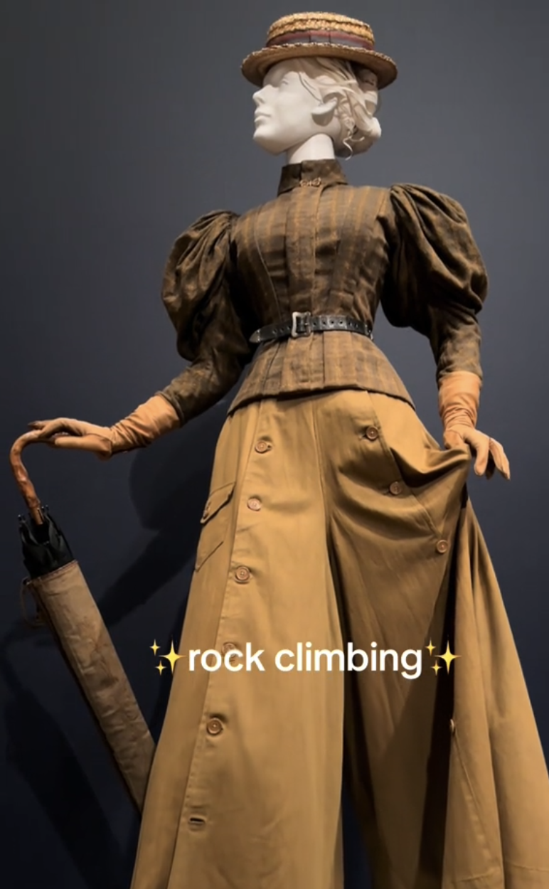 Mannequin dressed in early 20th-century outdoor attire with puffy-sleeve blouse, high-waisted pants, gloves, hat, and holding an umbrella, with text: &quot;rock climbing.&quot;