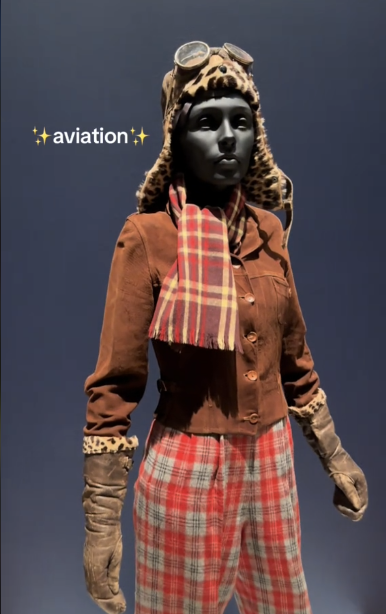 Mannequin dressed in vintage aviation gear with a leopard-print-lined helmet, goggles, brown jacket, gloves, red checkered scarf and pants under the word &quot;aviation.&quot;