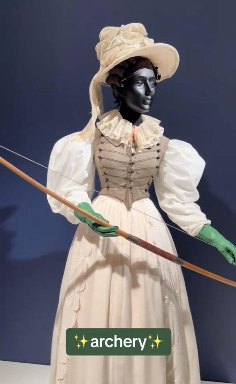A mannequin in a vintage-style white dress, green gloves, and a large hat holding a bow. Text reads &quot;✨archery✨&quot;