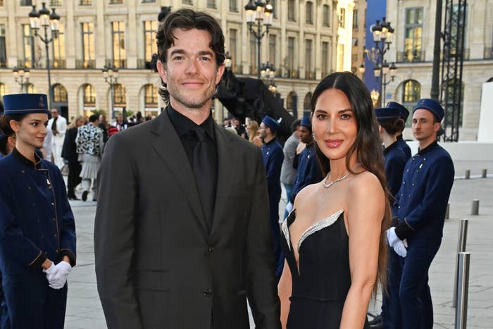 John Mulaney, in a black suit, and Olivia Munn, in a sleek black gown with an asymmetrical neckline, posing together at an outdoor event
