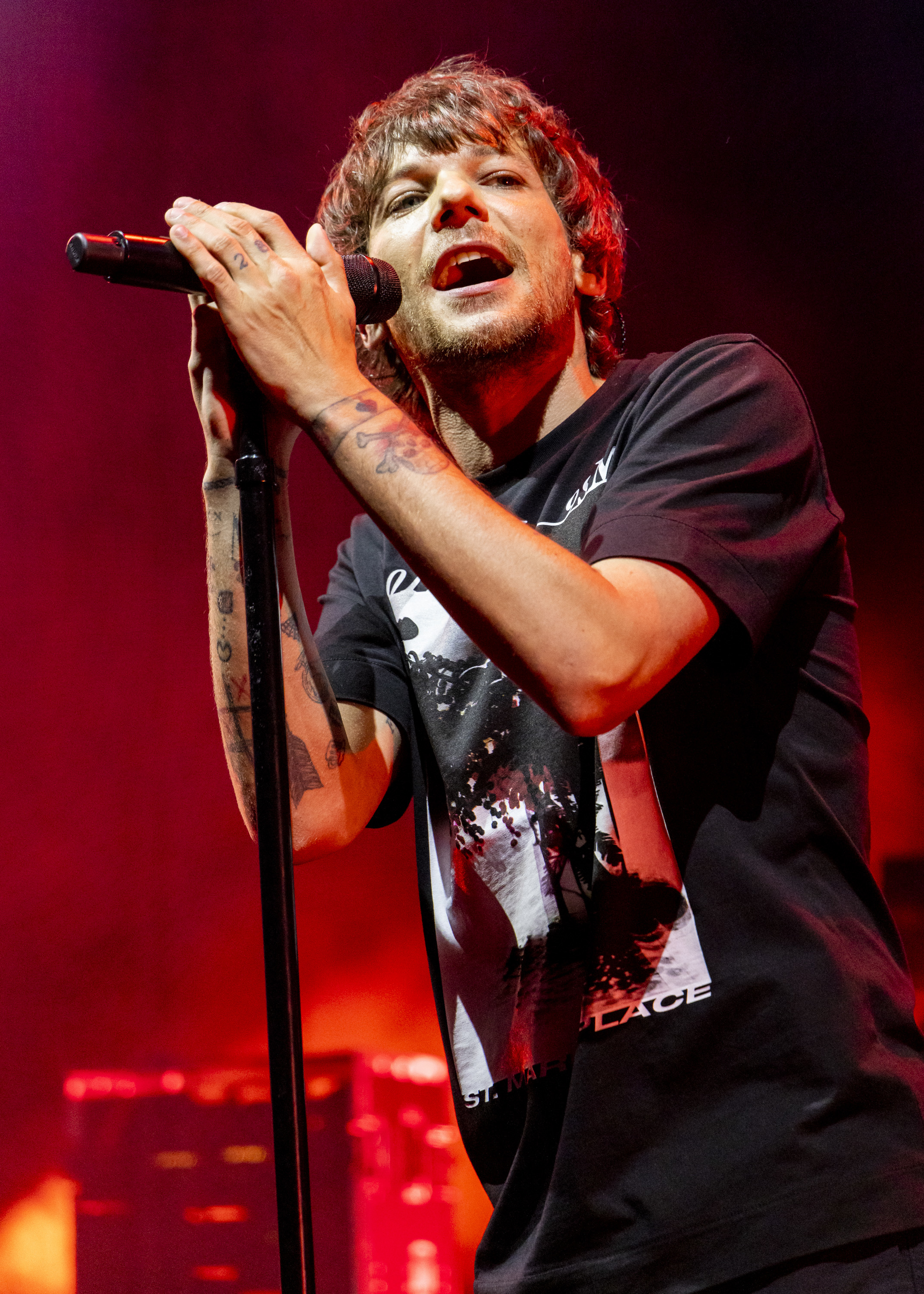 Louis Tomlinson performs on stage, singing into a microphone, wearing a graphic t-shirt
