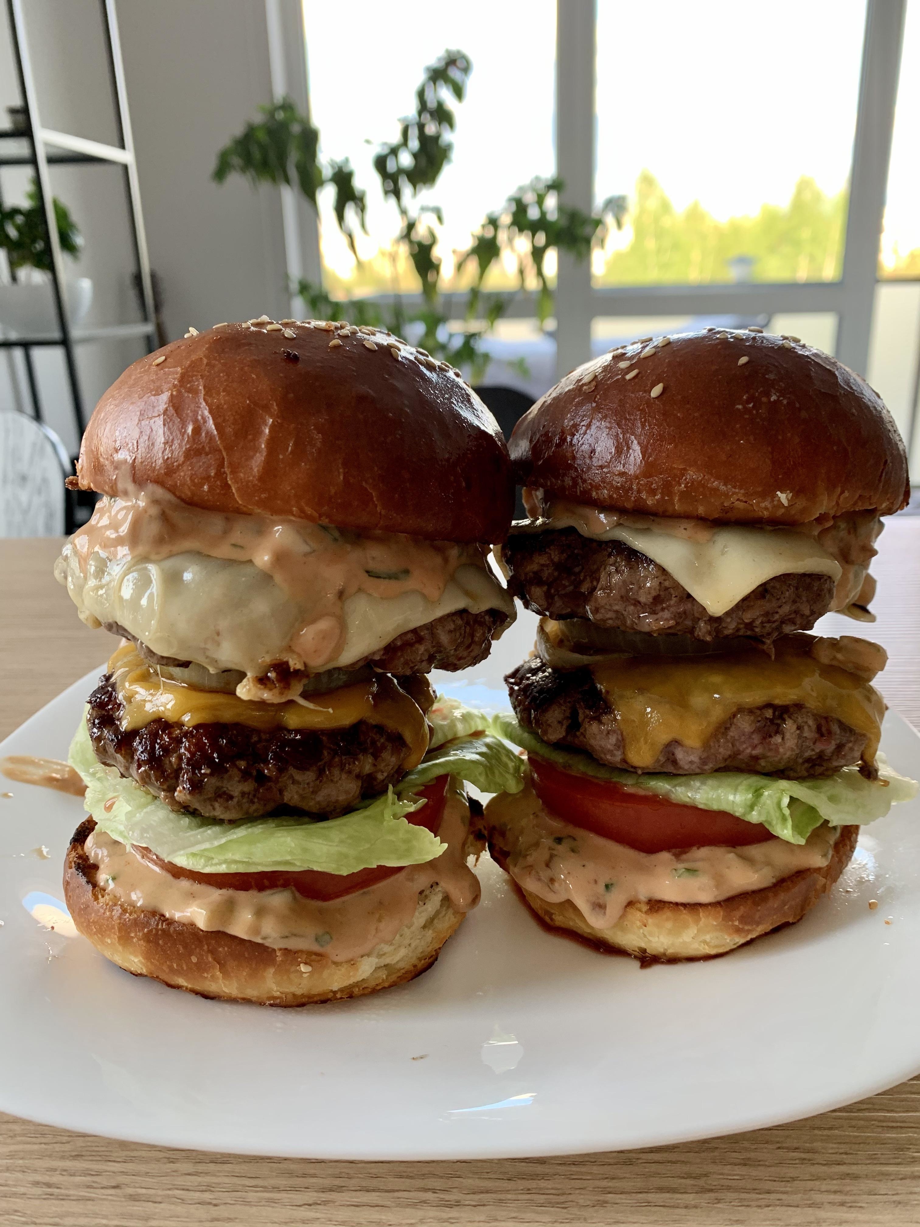 Two tall, homemade cheeseburgers stacked with lettuce, tomato, multiple beef patties, and various sauces on a white plate