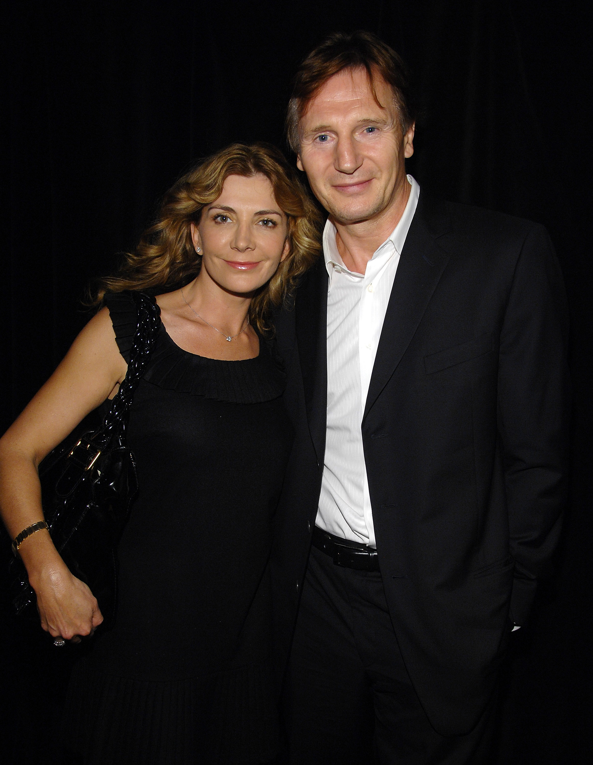 Natasha Richardson and Liam Neeson posing together, both in elegant black attire, with subtle smiles against a dark background